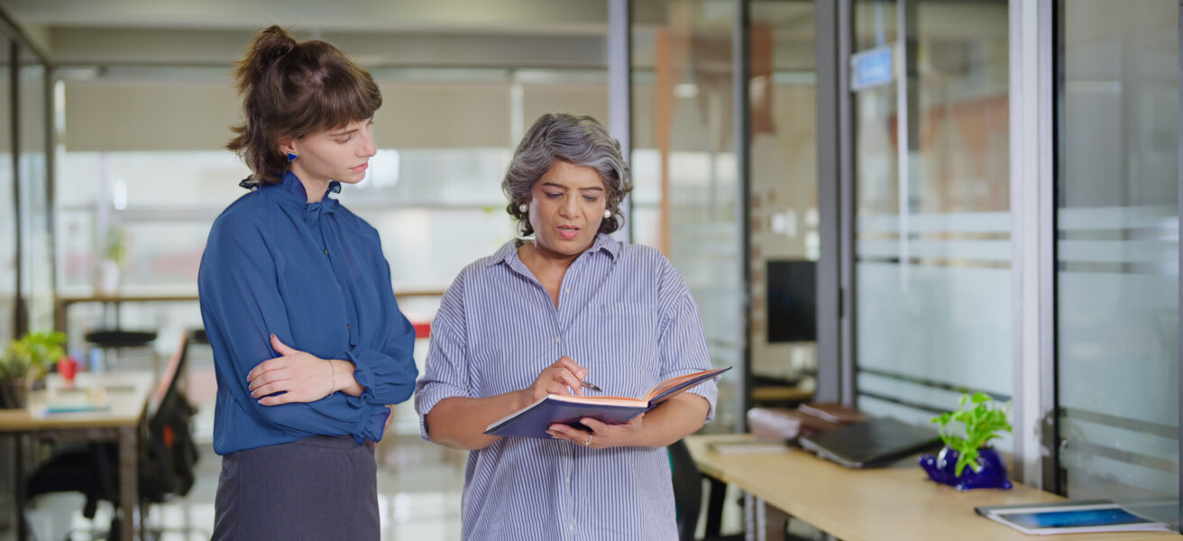 Indian senior old elder boss women lady standing hold book file make new project indoor office young adult expert intern girl older staff do work talk day task read report share ideas plan job place