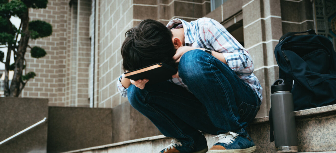 Good looking teenage Asian boy sit on stairway in front of high school building, bend his face down on notebook, frustrated, overwhelmed as he struggle for exam preparation, heavy homework, workload.