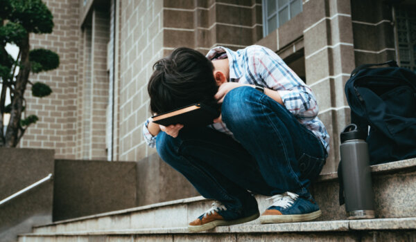 Good looking teenage Asian boy sit on stairway in front of high school building, bend his face down on notebook, frustrated, overwhelmed as he struggle for exam preparation, heavy homework, workload.